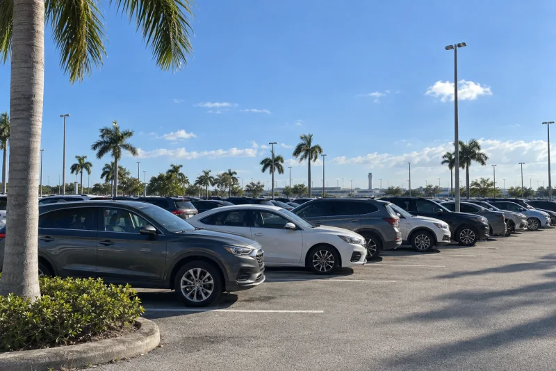 Rows of parked cars in an open-air economy lot with palms and a distant control tower